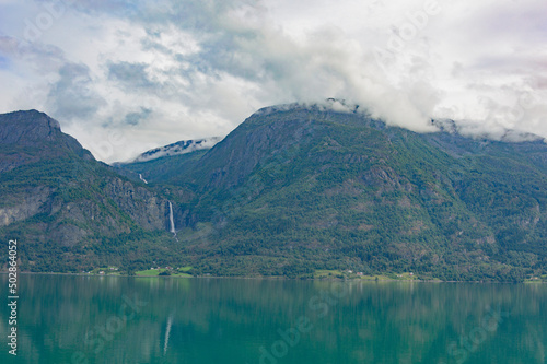 View of the Lustrafjord in Norway.  turquoise sea water  low white clouds above it  cloudy sky  green forest on the mountain slopes  white waterfall 