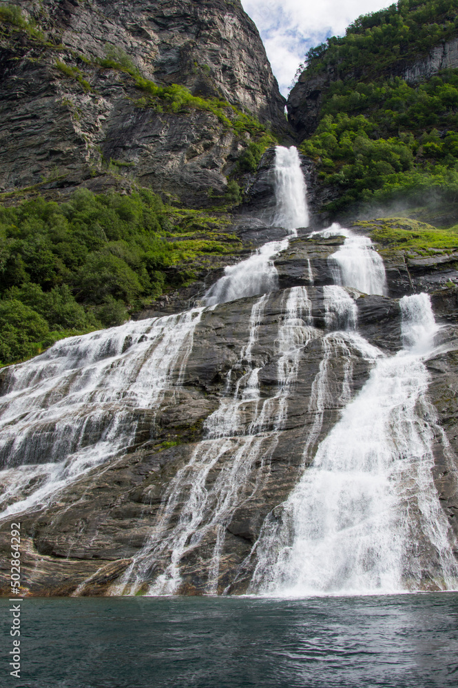 View of the Groom waterfall in Geirangerfjord, Norway from the water. Blue sky with clouds, green mounain slopes with sunshine and shadows on the forest View of the Groom waterfall in Geirangerfjord, Norway from the water. Blue sky with clouds, green mounain slopes with sunshine and shadows on the forest