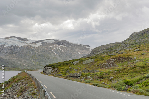road in the Norwegian mountains, summer landscape with snow. Green and grey rocky slopes, mossy stones, cloudy sky, grey water of the mountain lake