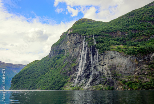 View of Geirangerfjord and the Seven sisters waterfall in Norway from the water.  Blue sky with clouds, green mounain slopes with sunshine and shadows on the forest