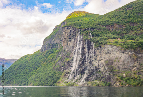 View of Geirangerfjord and the Seven sisters waterfall in Norway from the water. Blue sky with clouds, green mounain slopes with sunshine and shadows on the forest
