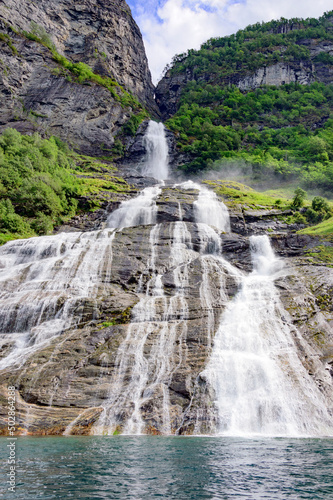 View of the Groom waterfall in Geirangerfjord  Norway from the water.  Blue sky with clouds  green mounain slopes with sunshine and shadows on the forest
