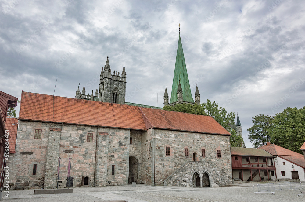 Nidarosdomen gothic cathedral and bishop`s palace  in Trondheim, Norway. Blue and lilac sky with grey clouds, grey medieval stone walls and bell towers,  green trees, cloudy sky
