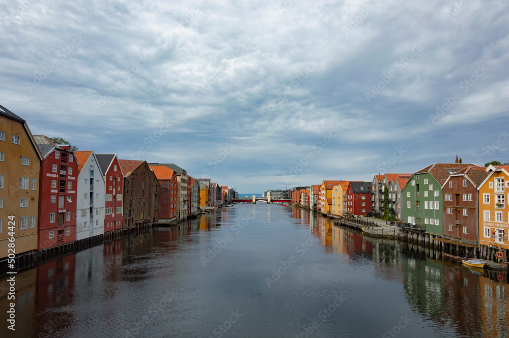 The Nidelva river flowing through the Trondheim city in Norway. Colourful old wooden storehouses on the river bank, grey water, cloudy sky