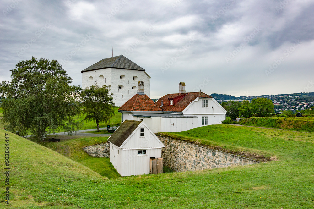 Citadel of the Kristiansten fortress in Trondheim, Norway. White tiny houses and walls with gun ports, green grass on the ramparts, grey cloudy sky