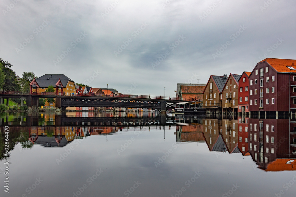 Fototapeta premium The Nidelva river flowing through the Trondheim city in Norway. Old wooden storehouses on the both river sides, old town bridge grey water, cloudy sky, 