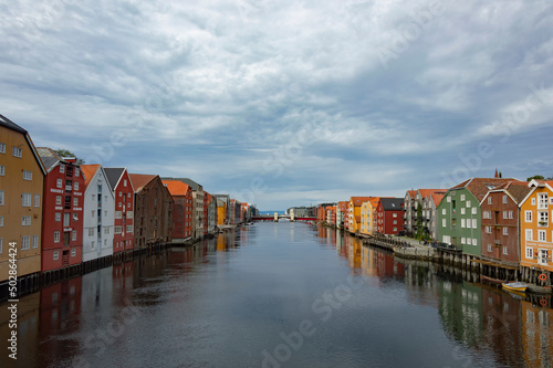 The Nidelva river flowing through the Trondheim city in Norway. Colourful old wooden storehouses on the river bank, grey water, cloudy sky