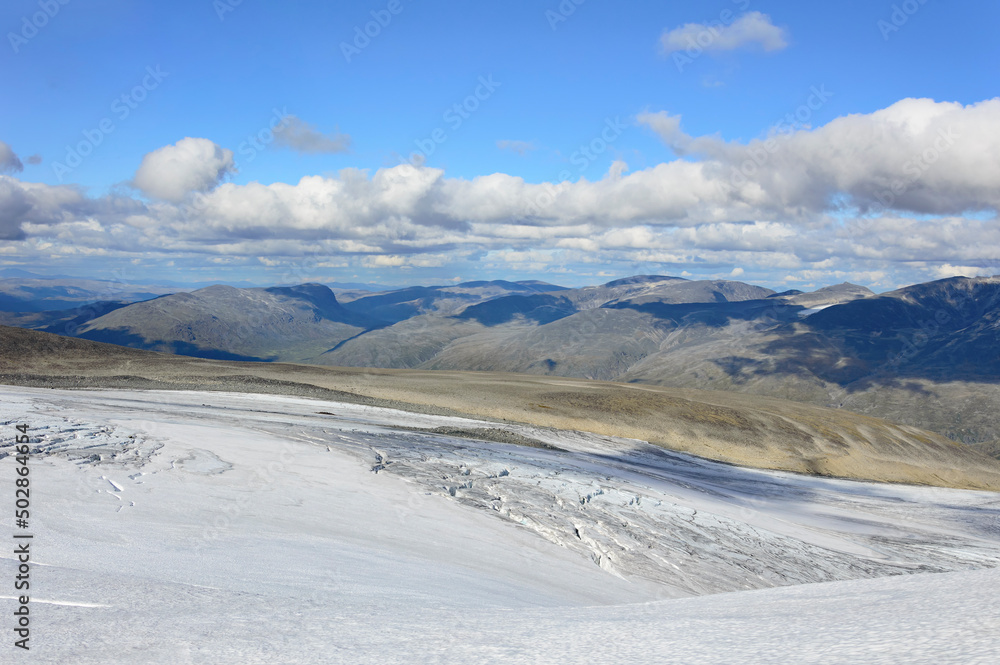 Wide panoramic view of the Jotunheimen national park in Norway from the Galdhopiggen mountain. Blue sky with clouds, grey and biege mountain slopes, white snow covered glacier Wide panoramic view of the Jotunheimen national park in Norway from the Galdhopiggen mountain. Blue sky with clouds, grey and biege mountain slopes, white snow covered glacier