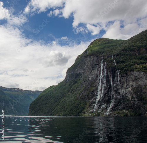 view of the cliff with the Seven Sisters waterfall from the water of Geirangerfjord in Norway. green forest on the rocky mountains around the fjord, blue sky with clouds