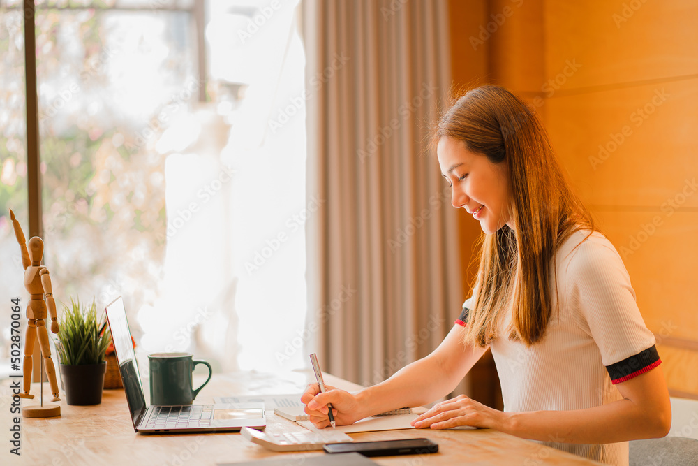 Cropped photo of beautiful Asian female accountant working from home checking documents and using notebooks and calculators. Saving at home smiling happily at work. Cropped photo of beautiful Asian female accountant working from home checking documents and using notebooks and calculators. Saving at home smiling happily at work.