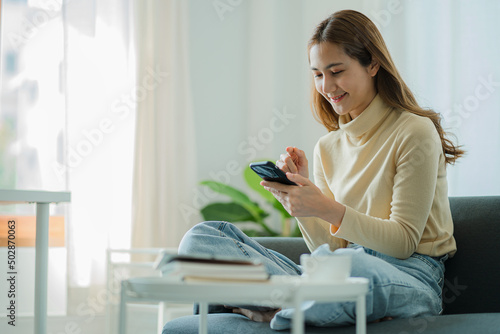 Asian woman using smartphone on sofa Use your mobile phone to surf the Internet, watch, play tech at home in the living room, relax, enjoy life.