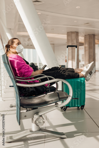 Vertical woman in mask with luggage sit in waiting room with smartphone at international airport terminal. Coronavirus