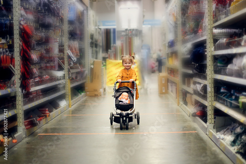 Cute baby girl in a mustard dress with doll in puppet stroller in a store, a child is playing in a hardware store