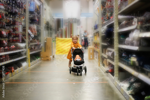 Cute baby girl in a mustard dress with doll in puppet stroller in a store, a child is playing in a hardware store