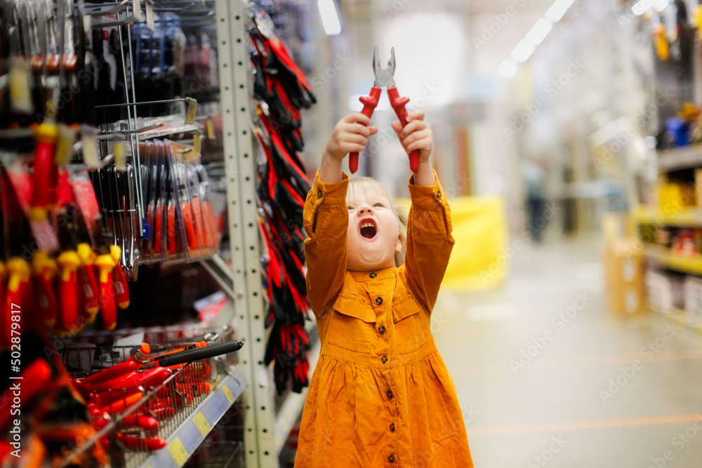 Cute girl child in mustard dress in hardware store, child plays with pliers, chooses tools in large hypermarket, child safety