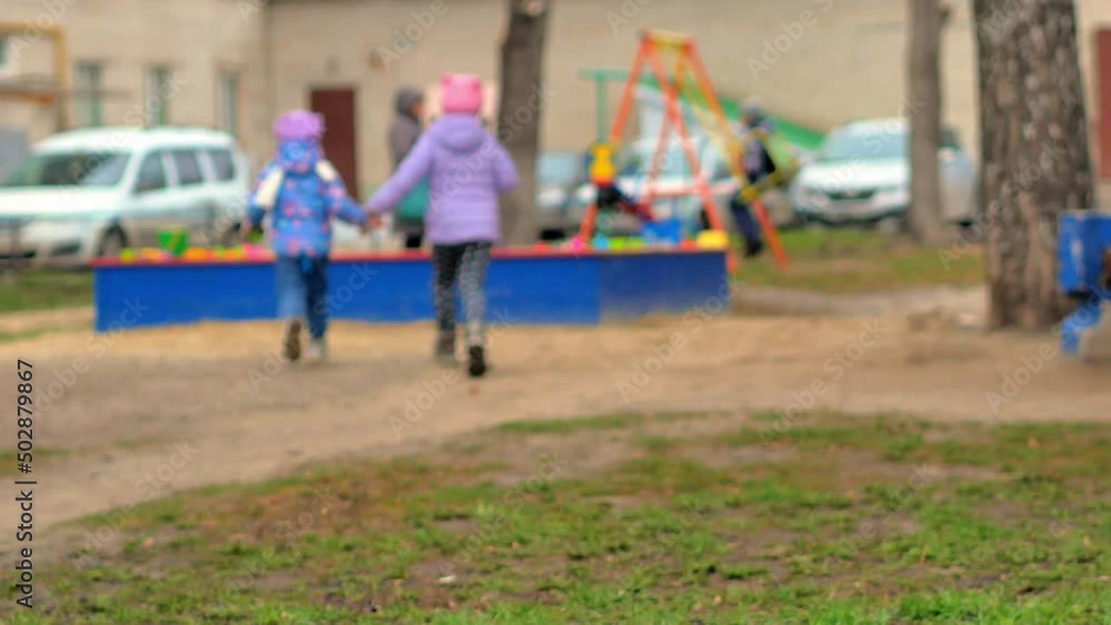 Funny little sister girls dressed in warm clothes in spring or autumn, holding hands, run together to the sandbox on the playground. The concept of sibling friendship. Selective focus. Funny little sister girls dressed in warm clothes in spring or autumn, holding hands, run together to the sandbox on the playground. The concept of sibling friendship. Selective focus.