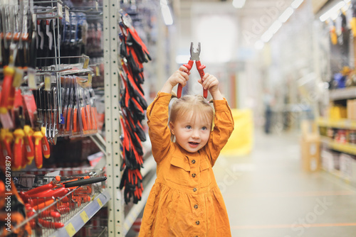 Cute girl child in mustard dress in hardware store, child plays with pliers, chooses tools in large hypermarket, child safety photo