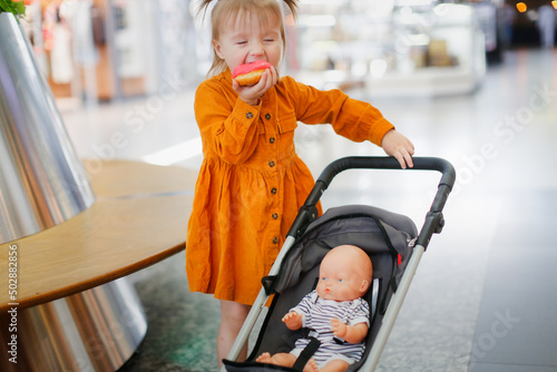 Toddler girl eats donut in supermarket on bench, cute child girl in mustard long-sleeve dress sits and feeds doll in stroller with donut. Girl playing with doll and stroller in store