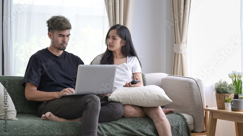 Happy young couple relaxing on couch in living room and using laptop together.