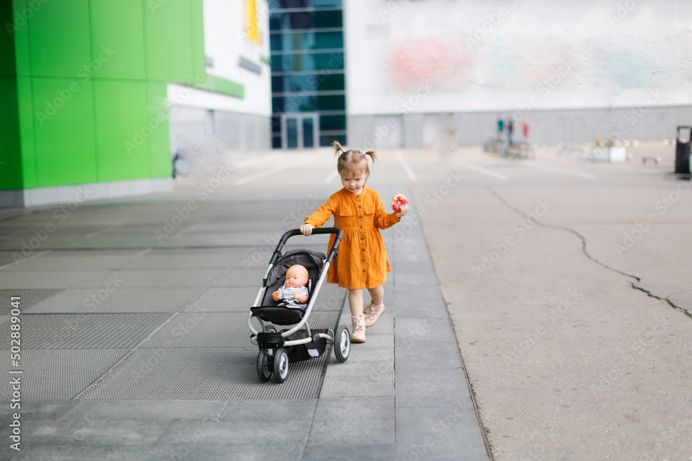 A cute toddler girl is walking with a stroller with a doll near the hypermarket. Caring and playing with a doll for a walk A cute toddler girl is walking with a stroller with a doll near the hypermarket. Caring and playing with a doll for a walk