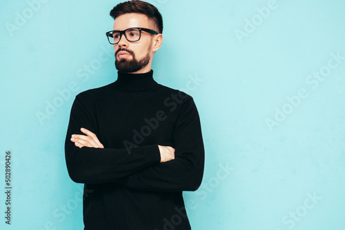 Handsome smiling model. Sexy stylish man dressed in black turtleneck sweater and jeans. Fashion hipster male posing near blue wall in studio. Isolated. In spectacles or eyeglasses