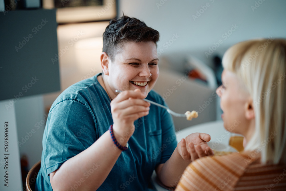 Happy gay woman sharing pasta with her girlfriend and feeding her during lunch at home. Happy gay woman sharing pasta with her girlfriend and feeding her during lunch at home.