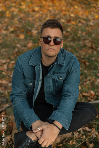 Handsome young man with stylish sunglasses in fashionable denim clothes with a blue jeans shirt sits in an autumn park and looks at the camera