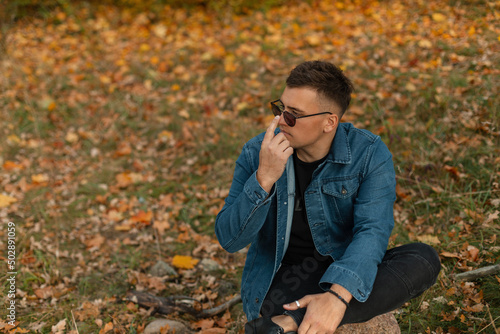 Fashionable handsome hipster man in a denim shirt with black clothes sits in nature with autumn yellow leaves and adjusts his sunglasses © alones