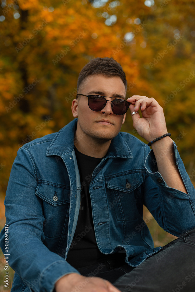 fashionable handsome young man model in trendy denim clothes sits in an autumn park and wearing vintage sunglasses. Fall male stylish look clothes fashionable handsome young man model in trendy denim clothes sits in an autumn park and wearing vintage sunglasses. Fall male stylish look clothes