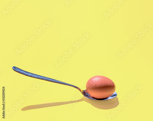 Egg and metal spoon on sunlight, with fancy shadow. Square composition on pastel yellow background, minimal abstract concept