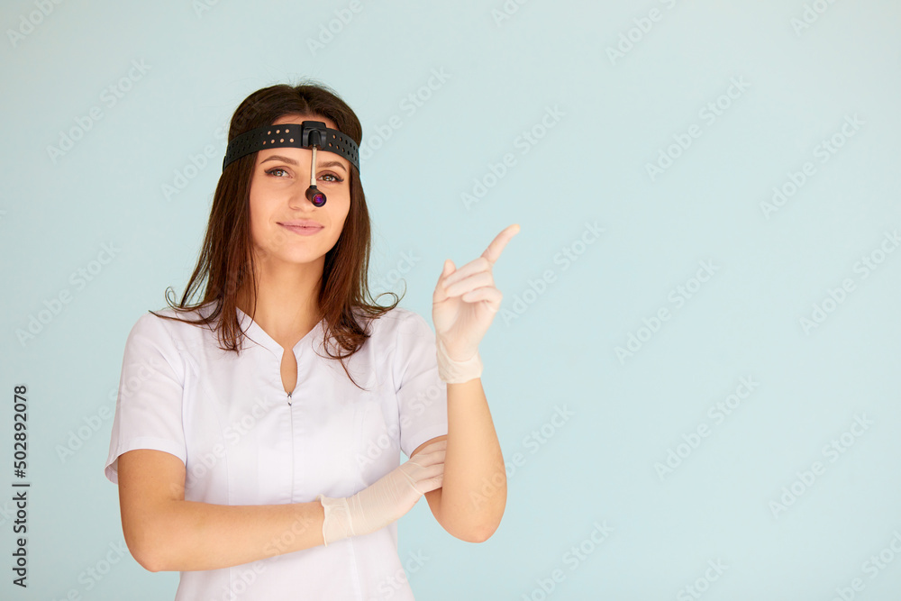 Cute young female otolaryngologist standing with illuminator on her head on bright background Cute young female otolaryngologist standing with illuminator on her head on bright background