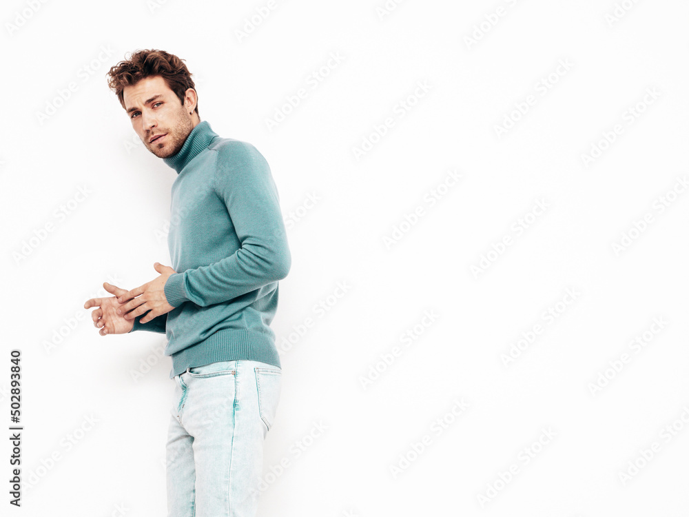 Portrait of handsome confident model. Sexy stylish man dressed in blue sweater and jeans. Fashion hipster male with curly hairstyle posing near white wall in studio. Isolated Portrait of handsome confident model. Sexy stylish man dressed in blue sweater and jeans. Fashion hipster male with curly hairstyle posing near white wall in studio. Isolated