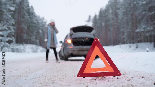 A woman walks around a broken car and calls from her smartphone. Winter, snow is falling. Breakdown on the road. auto insurance, call a tow truck.