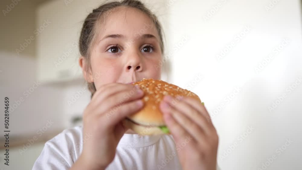 little girl eating a hamburger. unhealthy fast food proper nutrition concept. child greedily with pleasure bites a meal big burger in the kitchen at home. kid eats fast food close-up little girl eating a hamburger. unhealthy fast food proper nutrition concept. child greedily with pleasure bites a meal big burger in the kitchen at home. kid eats fast food close-up