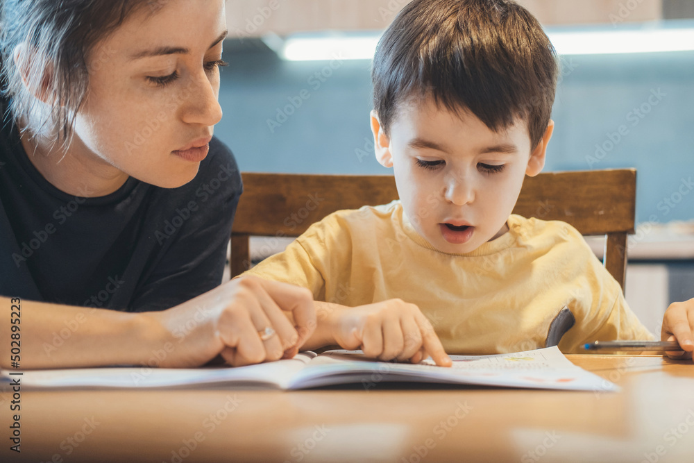 Caucasian little boy and his mother sitting together at table and reading book, mother teaching her child to read. Education, learning, study. Mother language. Caucasian little boy and his mother sitting together at table and reading book, mother teaching her child to read. Education, learning, study. Mother language.