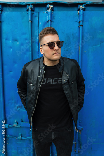 Fashion young handsome boy with black sunglasses in leather jacket, jeans and black t-shirt stands near a metal blue background