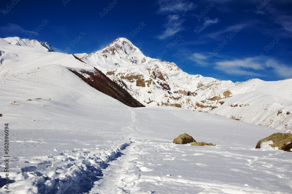 Hiking trail in the mountains. Landscape with snow-capped mountain peaks and clouds in the blue sky. The footpath leads to the foot of the Caucasus Mountains. Caucasian mountains