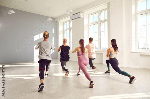 Group of young people exercising together. Team of men and women in sportswear doing forward lunges for their leg muscles during a fitness workout at the gym. Back rear backside view from behind
