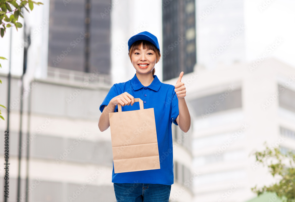 service and job concept - happy smiling delivery woman in blue uniform with takeaway food in paper bag showing thumbs up over city street background service and job concept - happy smiling delivery woman in blue uniform with takeaway food in paper bag showing thumbs up over city street background