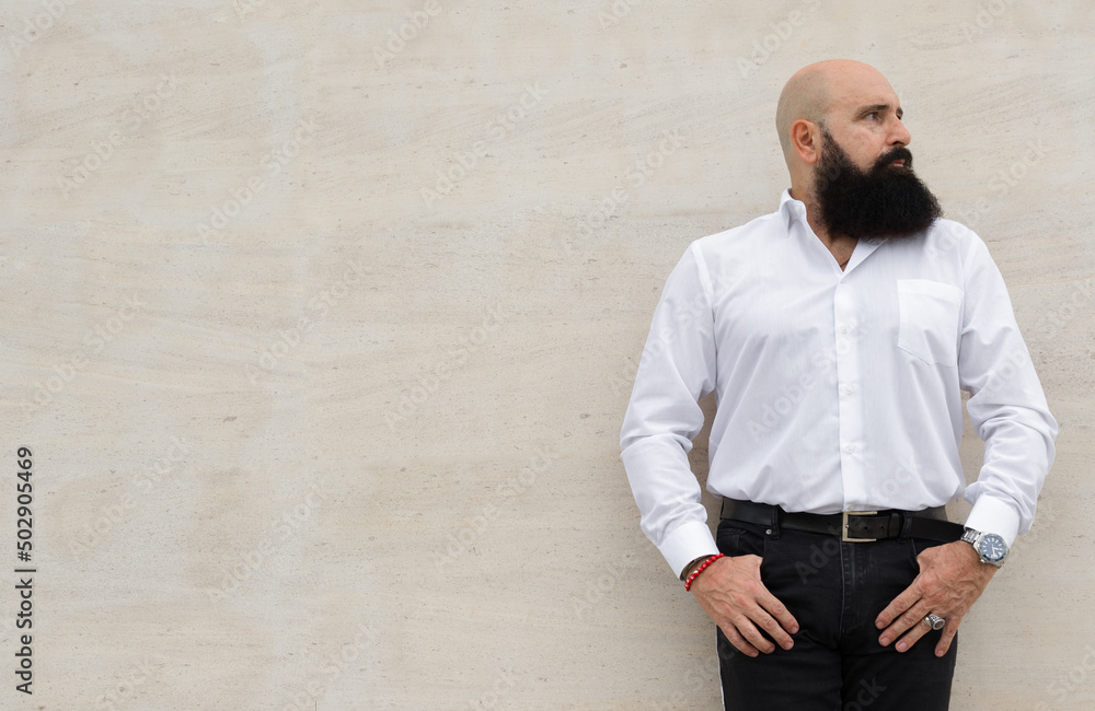 Shot of a hairless head man with a cared black beard that is leaning on a unicolor wall while have his hands in the pockets and is looking to the left Shot of a hairless head man with a cared black beard that is leaning on a unicolor wall while have his hands in the pockets and is looking to the left