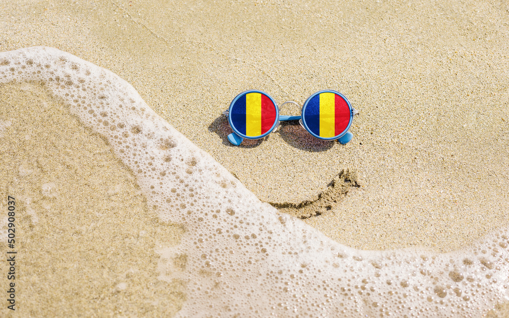 Obraz premium Sunglasses with flag of Romania on a sandy beach. Nearby is a sea lightning and a painted smile. The concept of a successful vacation in the resorts of Romania.