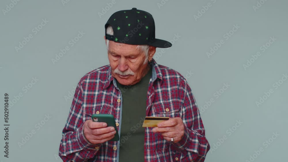 Sincere mature man customer using credit bank card and smartphone while transferring money, purchases online shopping, payment. Finance and internet. Senior grandfather alone on gray wall background Sincere mature man customer using credit bank card and smartphone while transferring money, purchases online shopping, payment. Finance and internet. Senior grandfather alone on gray wall background