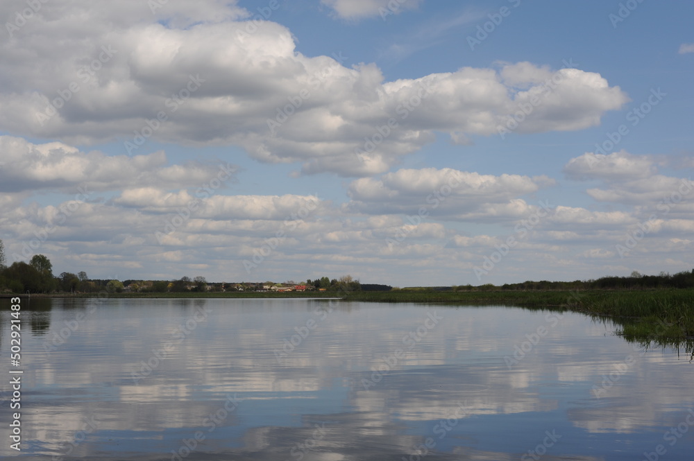 Fototapeta premium Picturesque and peaceful cloudscape with white clouds and blue sky reflecting in the river in spring or summer in Poland