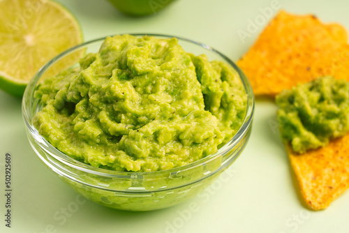 Close up view of mexican dip, spread or salad made of pureed ripe avocado served in glass bowl with crispy tortilla chips or nachos and halved lime slices as snack or appetizer on green background