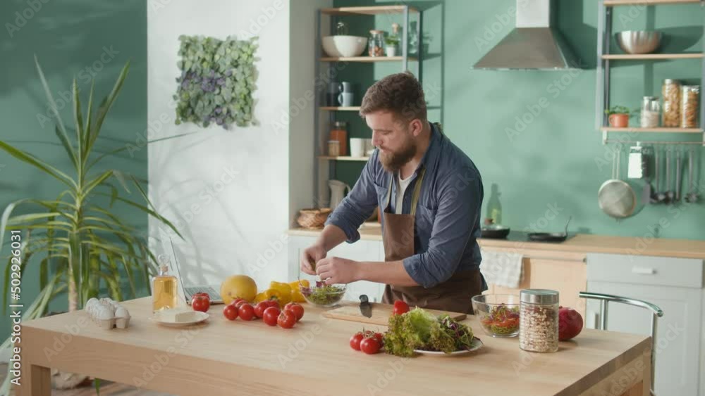 Bearded Man Cutting Tomato on Cutting Board in Kitchen, Checking for Recept in Laptop. Handsome Young Man Cooking Vegetarian Breakfast Cutting Vegetables for Salad in Kitchen at Home.Healthy Lifestyle Bearded Man Cutting Tomato on Cutting Board in Kitchen, Checking for Recept in Laptop. Handsome Young Man Cooking Vegetarian Breakfast Cutting Vegetables for Salad in Kitchen at Home.Healthy Lifestyle