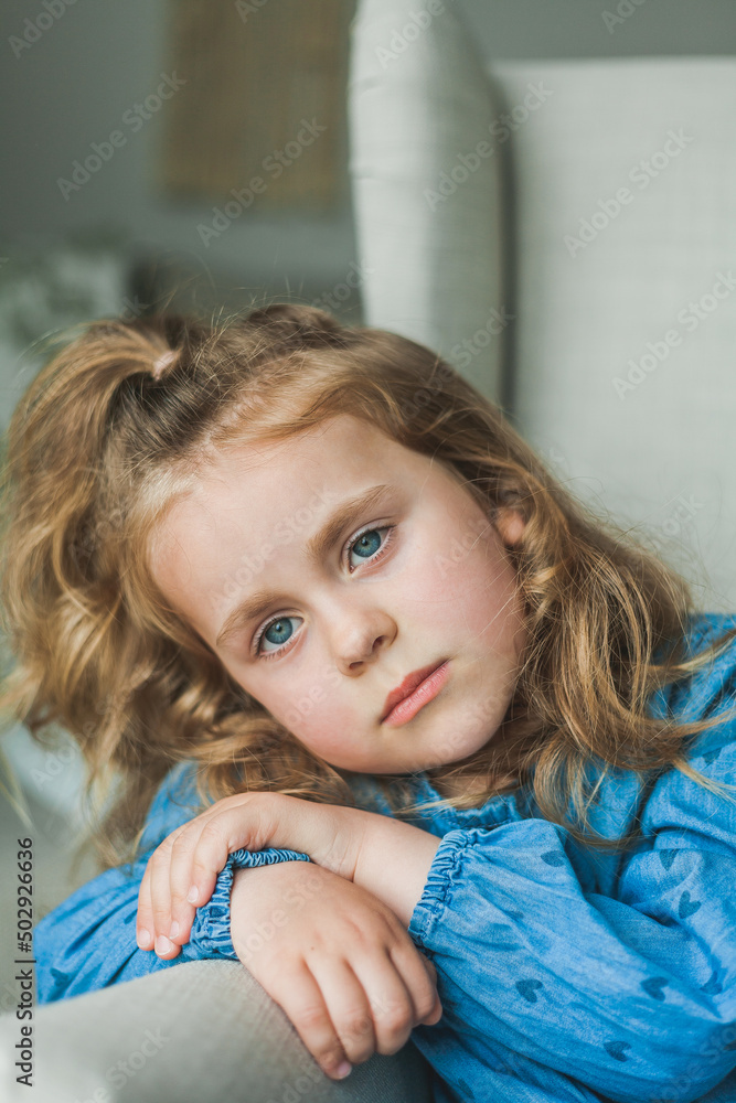 A little cute blue-eyed girl with curly long hair sits in a cozy armchair at home. Cozy, bright house. Happy child is resting. A little cute blue-eyed girl with curly long hair sits in a cozy armchair at home. Cozy, bright house. Happy child is resting.