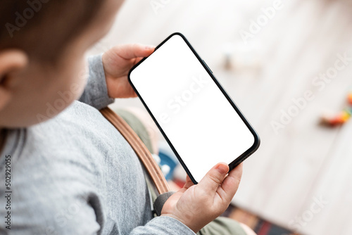 Boy's toddler hands holdingphone on home background. Child's hands hold black smartphone and watching video in horizontal position. Screen blank. Close-Up. photo