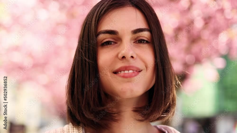 Portrait of a cute dark haired woman smiling charmingly while standing in the city street with sakura flowers in the background. Beautiful woman looking at camera outside. Portrait of a cute dark haired woman smiling charmingly while standing in the city street with sakura flowers in the background. Beautiful woman looking at camera outside.