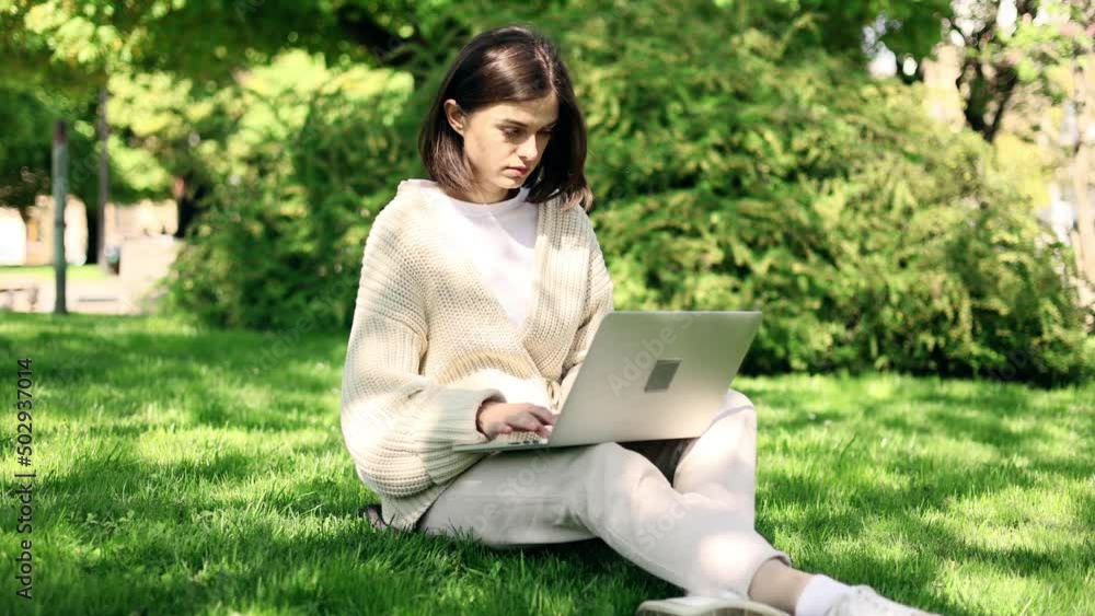 Attractive dark haired woman working at the laptop as sitting on grass in city park. Freelancer working in the park. Student study outside. Attractive dark haired woman working at the laptop as sitting on grass in city park. Freelancer working in the park. Student study outside.