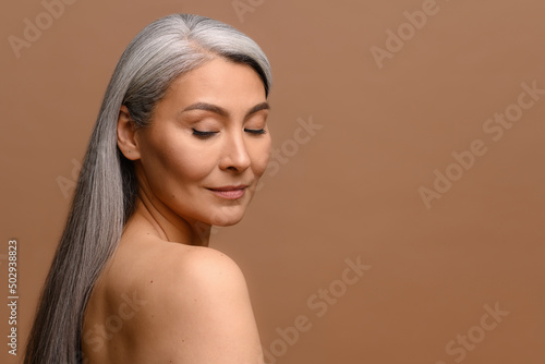 Headshot of attractive middle-aged Asian woman with naked shoulders and long silver hair isolated on brown, fascinating mature korean lady with smooth skin looks down dreamily. Skincare, copy space photo
